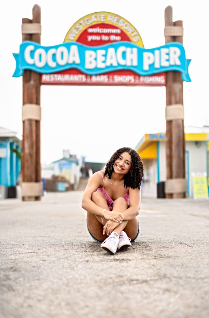 Senior photo of girl sitting and smiling under the inconic Cocoa Beach Pier sign with confidence.