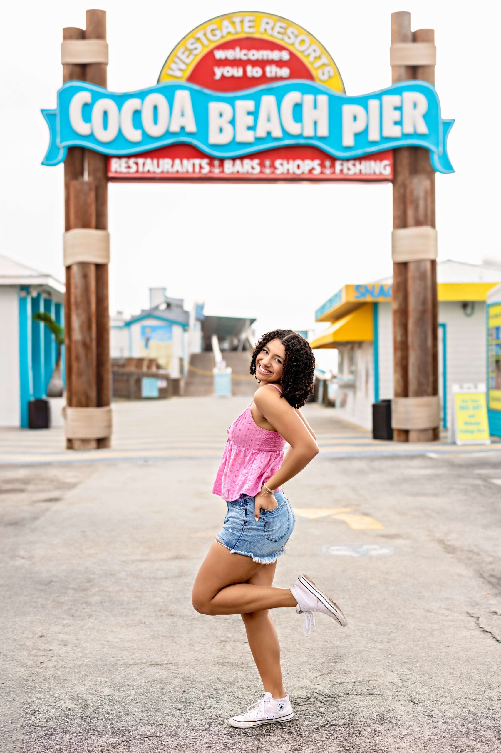 High school senior girl smiling and lifting one leg in front of the colorful Cocoa Beach Pier sign during her sunrise photo session in Central Florida