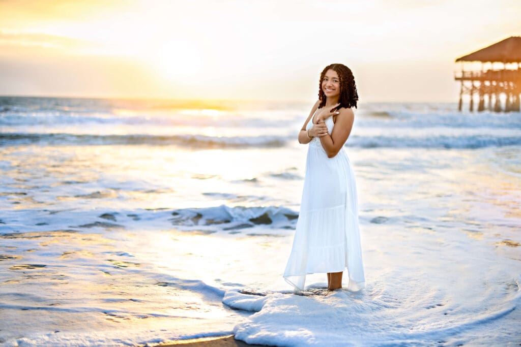 Teen girl standing in foamy waves at sunrise with soft golden light and ocean reflecting around her at Cocoa Beach.