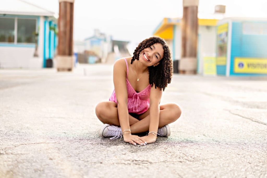 Teen girl sitting cross-legged in the middle of the road under the Cocoa Beach Pier sign, smiling big with beach buildings in the background.