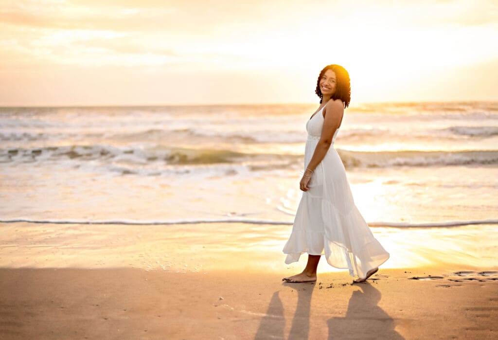 Senior girl walking barefoot along the ocean's edge during golden sunrise in a gorgeous white dress at Cocoa Beach.