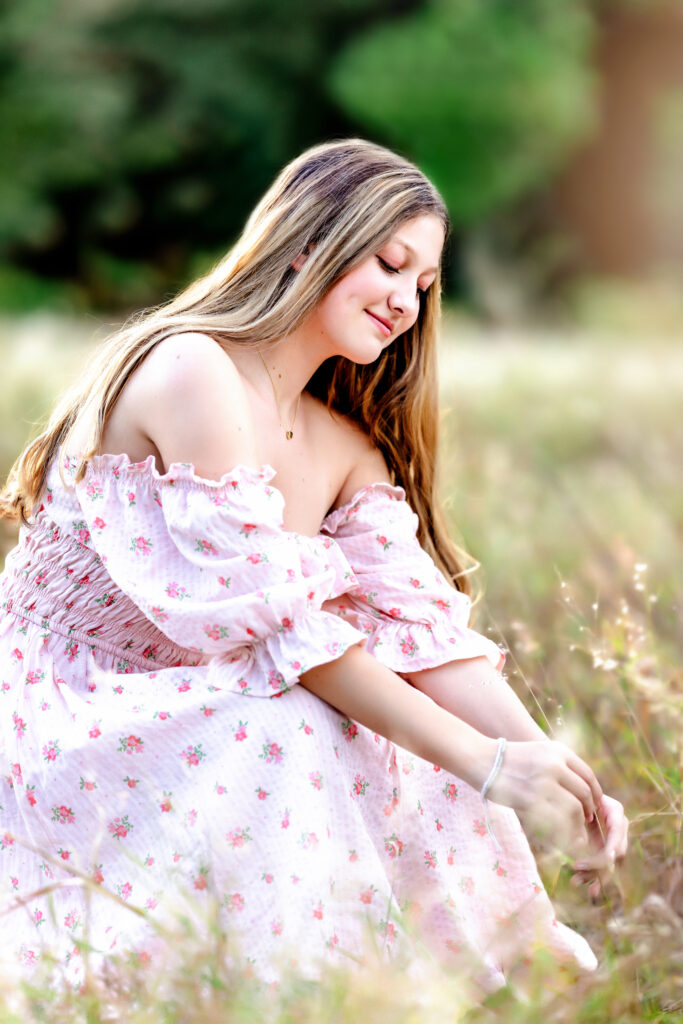 Teen girl seated in sunlit field, smiling while looking down during her senior session in Orlando, Florida.