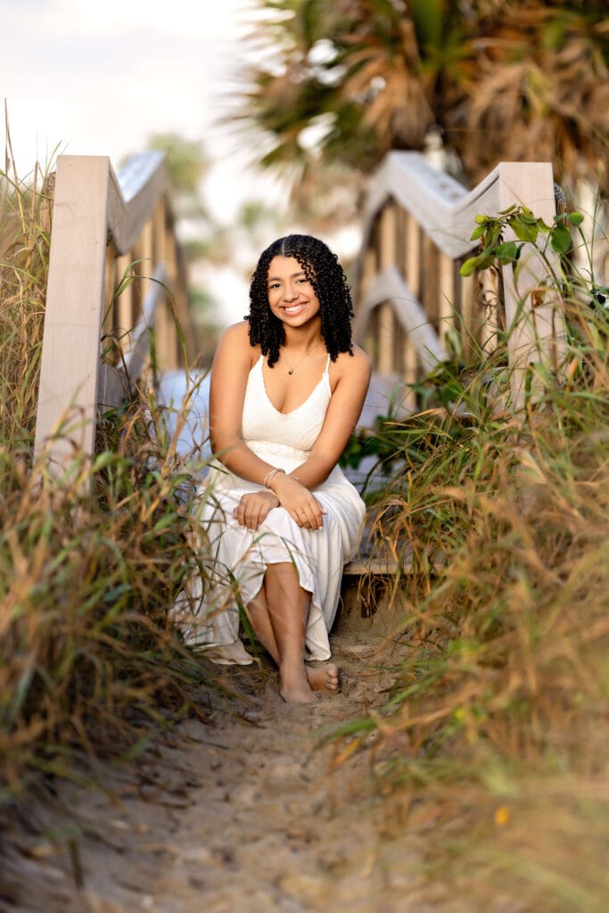 Teen girl sitting barefoot in golden sand with beach grass around her, glowing from beautiful light.