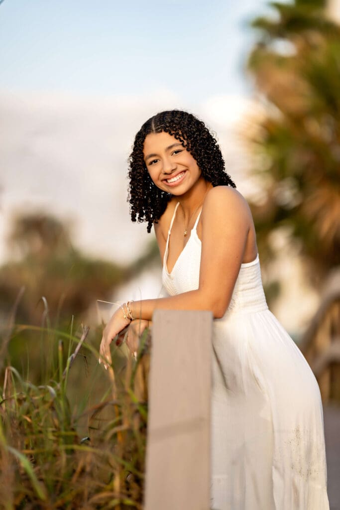 Senior girl in white dress leaning against a wooden railing with soft beach grass and palm trees in the background near Cocoa Beach.