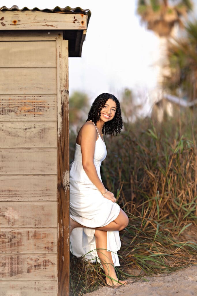 Senior photos of teen girl leaning up against a rustic wooden beach shack with a bright smile, curly hair, and white dress near Cocoa Beach Florida.