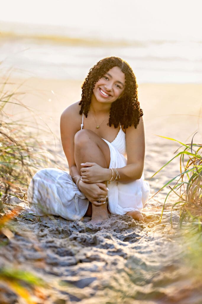 Teen girl sitting barefoot in golden sand with beach grass around her glowing with golden light.