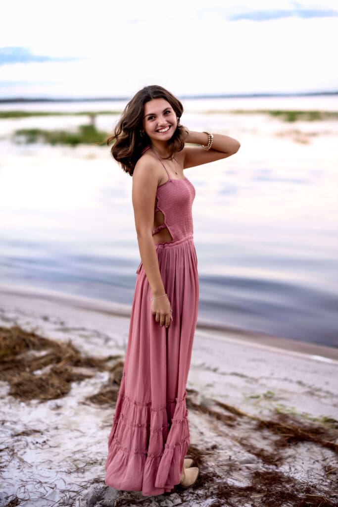 High school girl standing next to a lake right after a storm had rolled through. The skies are moody but she is radiant in a pink dress with windblown hair. 