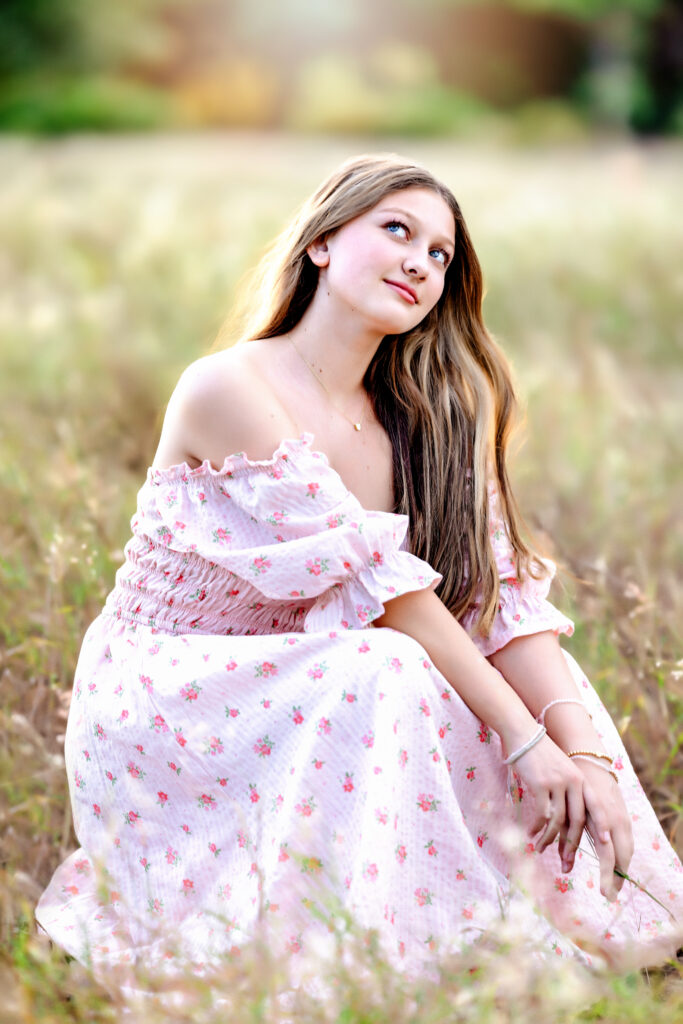 Senior girl wearing a floral off-the-shoulder dress smiling in a glowing field during golden hour, photographed by a Central Florida senior photographer in Windermere.