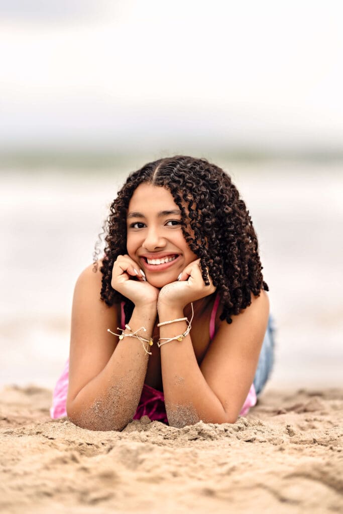 Teen girl lying on her stomach in the sand, hands on chin, smiling in front of the shoreline.