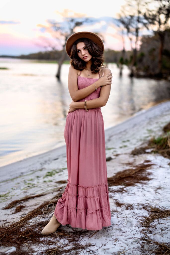 Teen girl in a blush pink maxi dress and wide-brim hat posing by the lakeside at Lake Louisa State Park, one of the best senior photo locations near Orlando, Florida. 