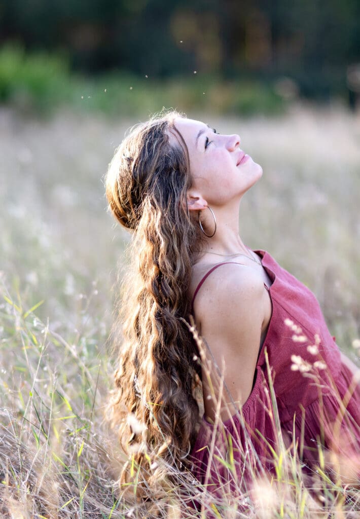 High school senior girl sitting in a tall grass field during golden hour in Orlando, Florida, smiling uo into the sun as tiny gnats swirl around her hair, effortlessly slaying her senior photos despite the full airborne cast of A Bug's Life. 