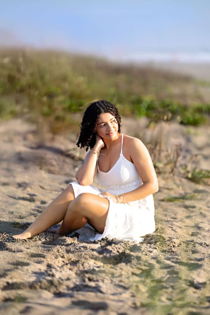 Teen girl sitting barefoot in the sand with beach grass and ocean waves surrounding her in the distance smiling in direct sunlight.