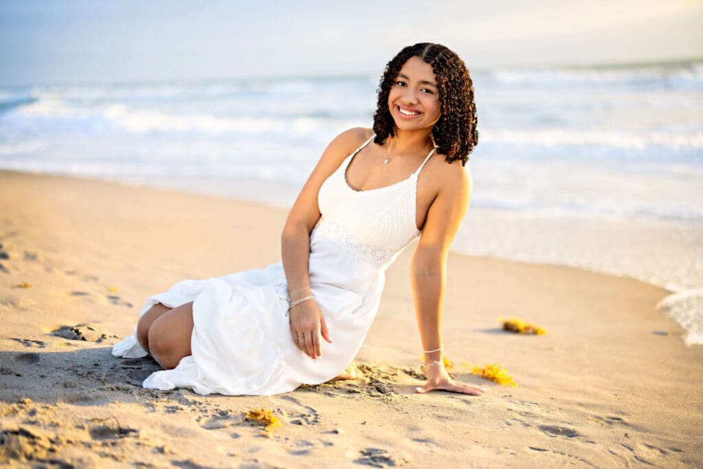 Teen girl in a white dress sitting on the beach near the tide line, surrounded by seaweed at Cocoa Beach Pier during sunrise.
