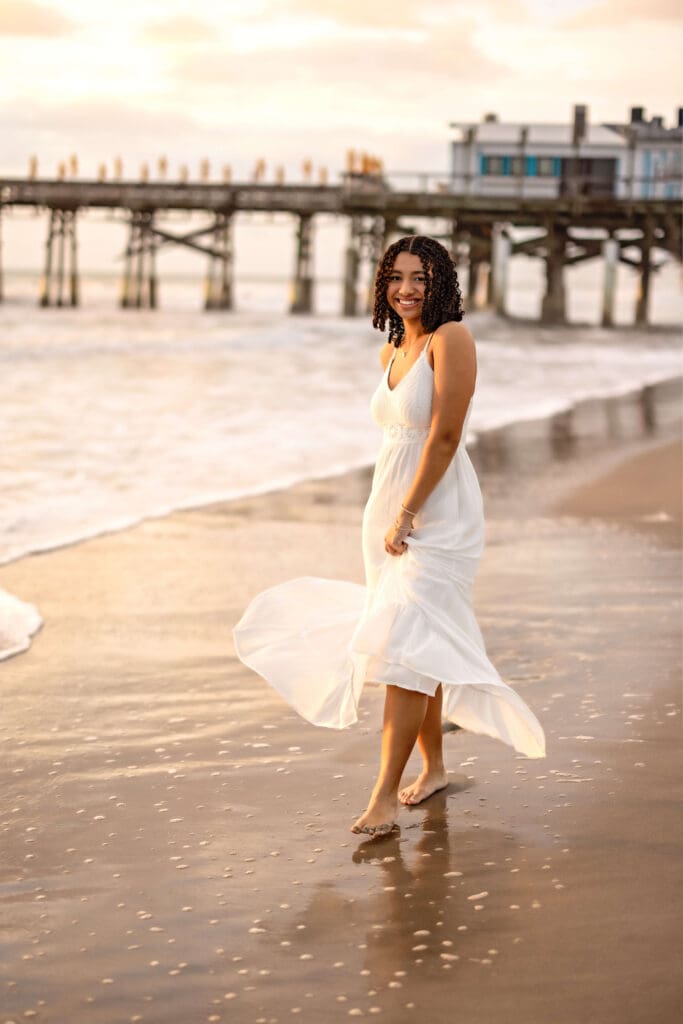Senior girl twirling barefoot in the sand under the Cocoa Beach Pier with wind catching her white dress in a very cinematic way.