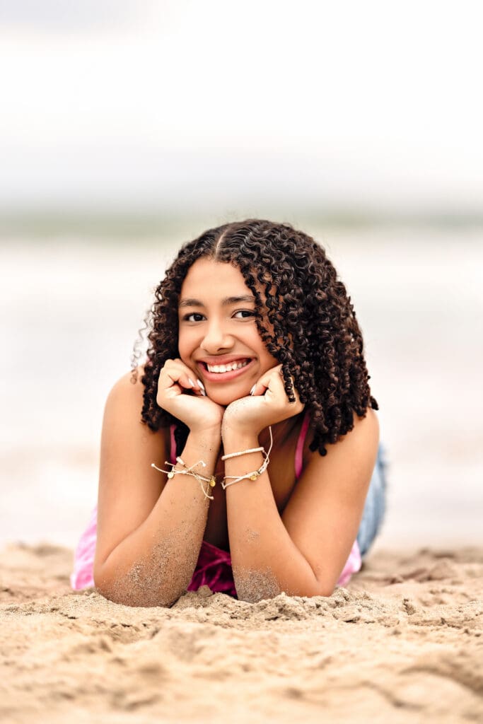 Senior girl sitting in the sand at Cocoa Beach, smiling softly and wearing layered Pura Vida bracelets, showing how accessories can elevate a senior photo outfit during a Florida beach senior session.