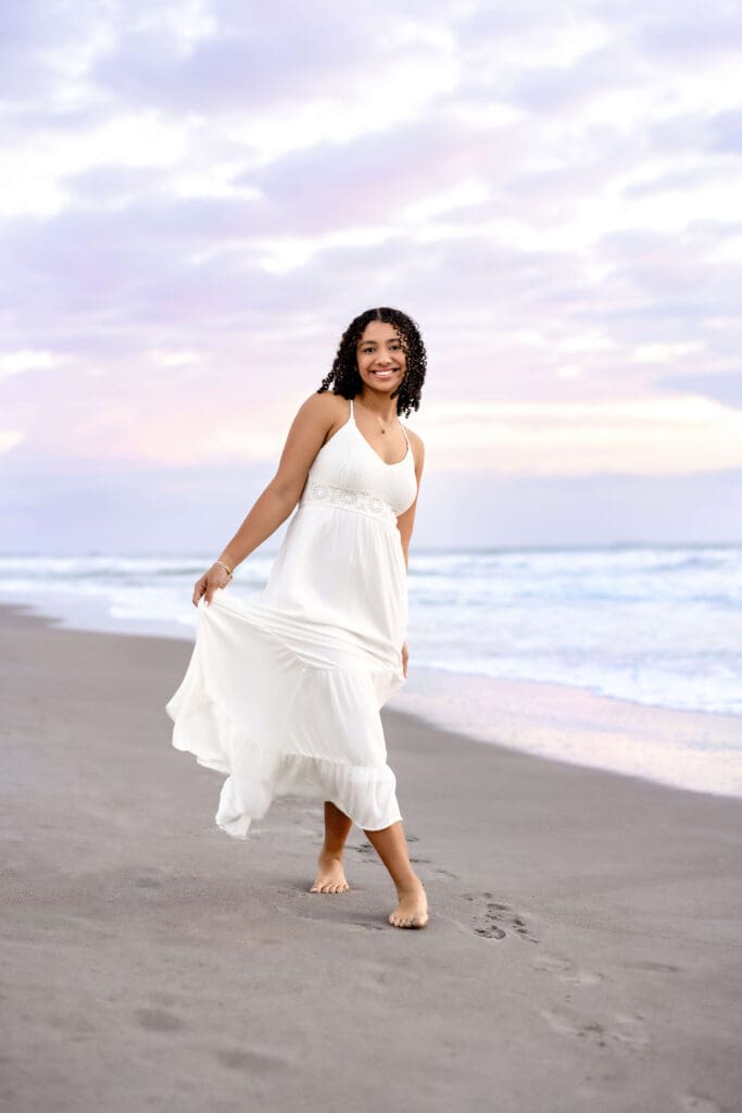 Senior girl walking barefoot along the shore in a flowing white dress with pastel clouds behind her at Cocoa Beach.