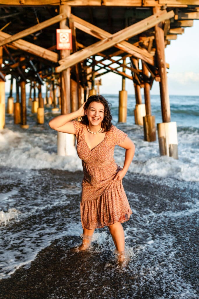 Senior girl in a rust-colored dress standing under the Cocoa Beach Pier as ocean waves crash around her feet, golden hour light hitting the wooden beams.