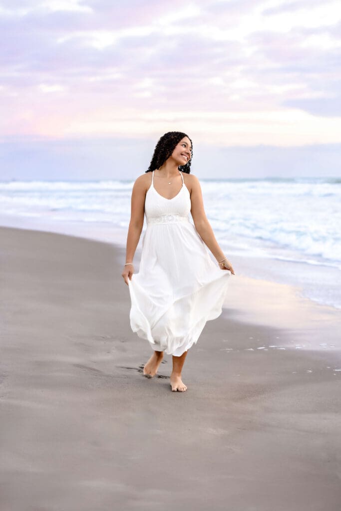 Senior girl in a flowing white dress walking. barefoot along Cocoa Beach during sunrise, soft pastel sky and gentle waves in the background. 