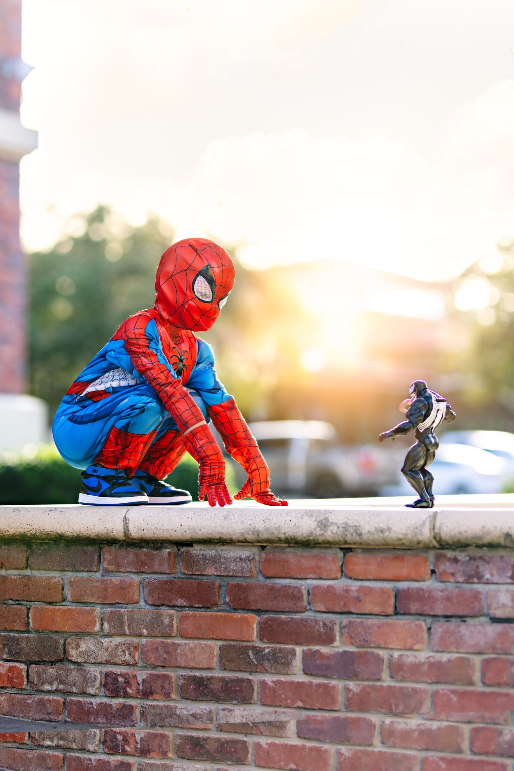 Young boy in a Spider-Man costume crouched on a brick ledge in Downtown Winter Garden, facing off with a Venom toy during a cinematic-inspired photo shoot.