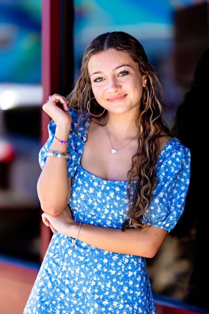 Senior girl in a blue floral dress posing with soft smile and arm raised beside a glass storefront reflecting downtown textures in Orlando.