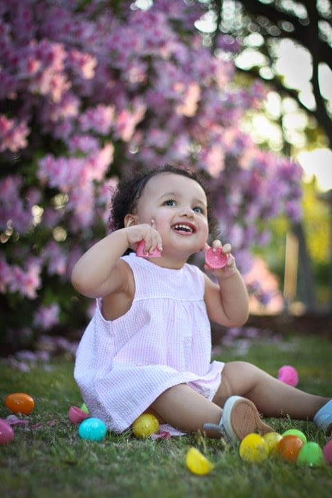 Toddler girl in a pink dress sitting in grass surrounded by plastic Easter Eggs and flowers, smiling at the camera with natural light.