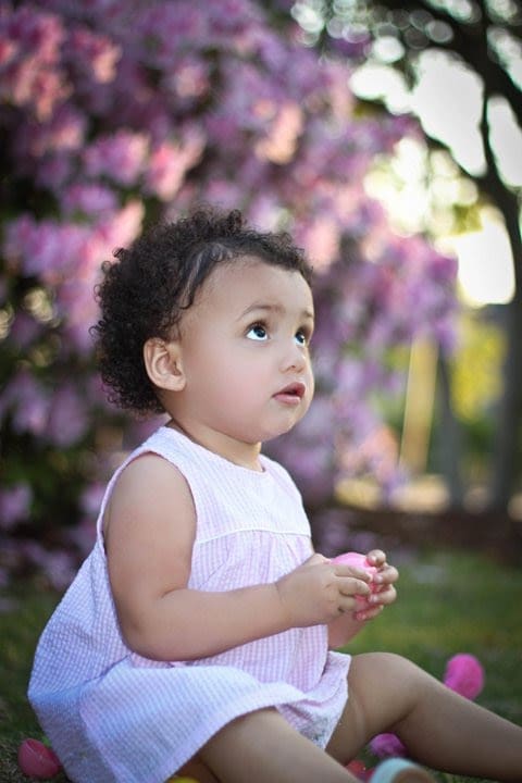 Toddler girl in a smocked Easter dress playing with pink egg and looking up sweetly while seated on grass. 