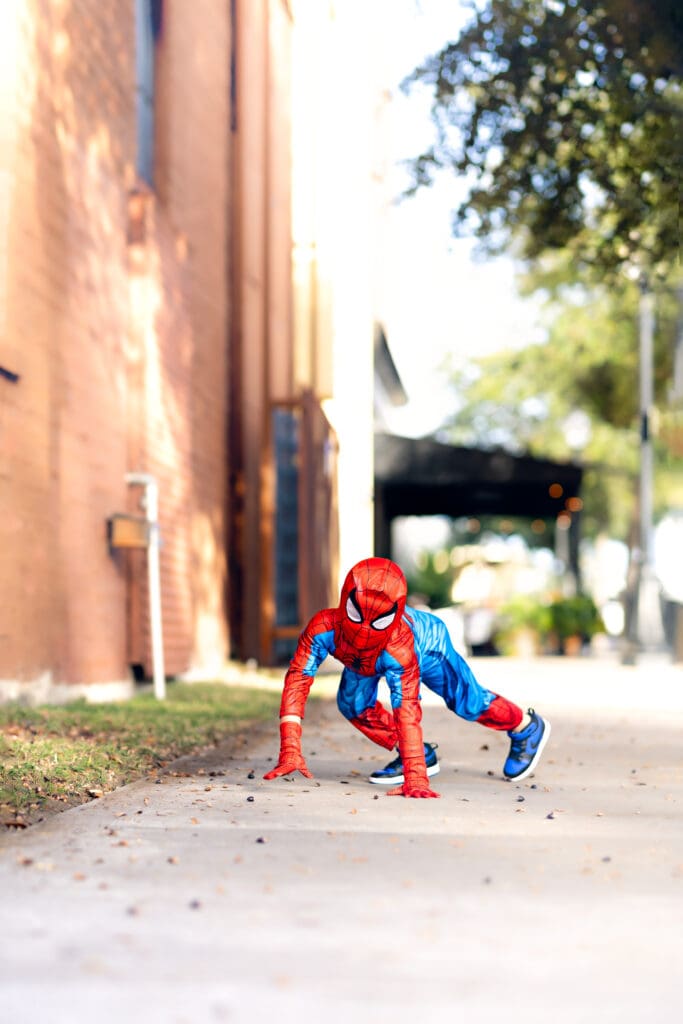Boy in a Spider-Man costume crouched low on all fours on a Downtown Winter Garden sidewalk, mid-hero pose during a creative shoot from senior photographer mom.