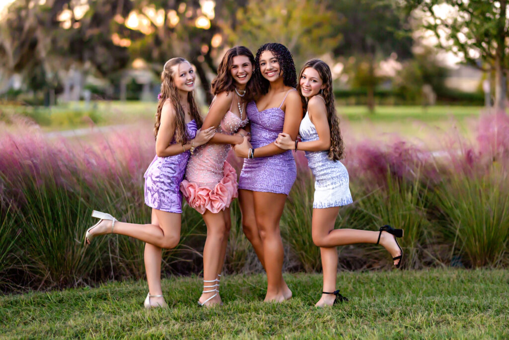 Four high school girls in sparkly homecoming dresses posing together in a field of pink muhly grass during sunset in Winter Garden, Florida.