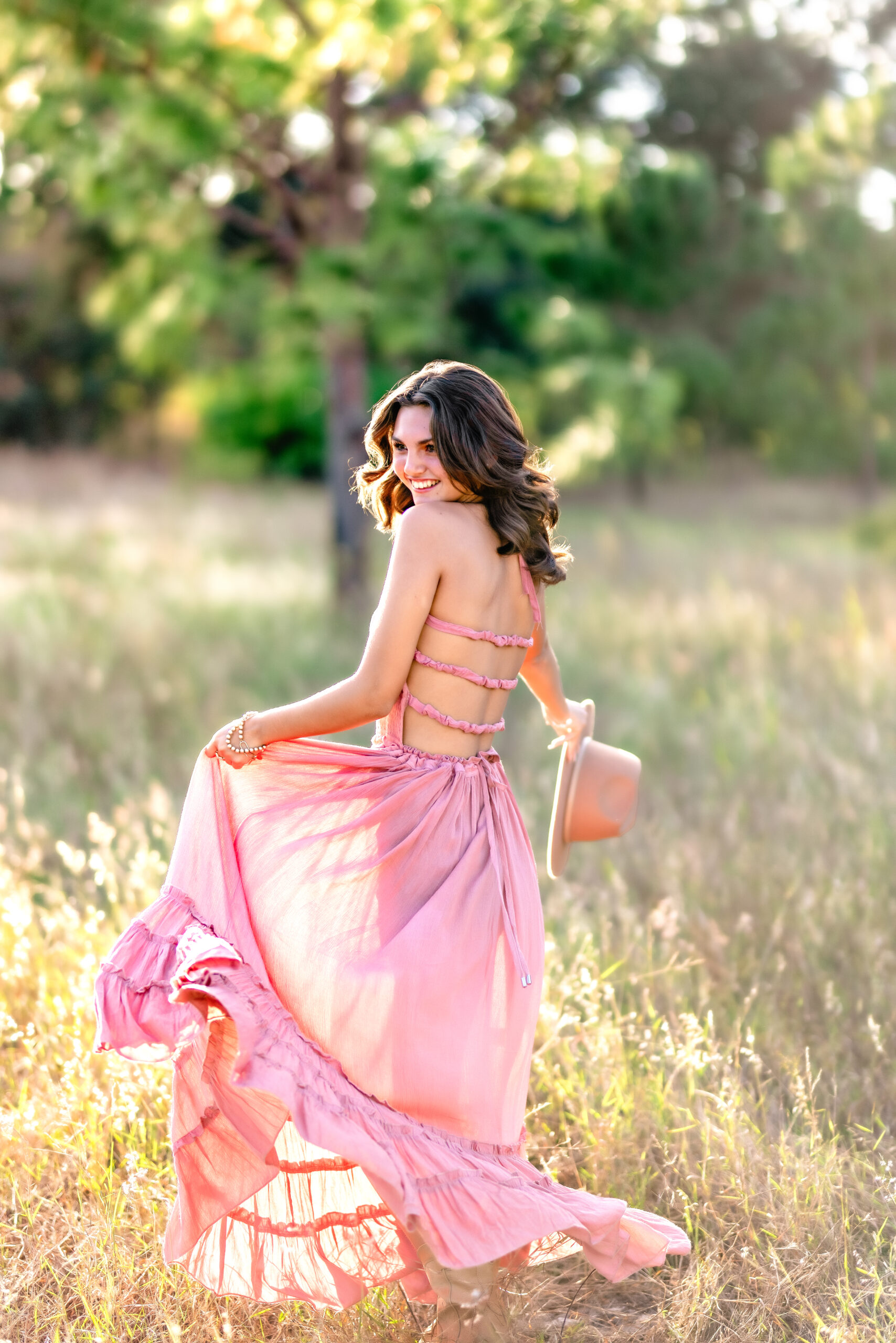 High school senior in a pink dress spinning in a sunlit field, holding a wide-brimmed hat during her senior photo session in Orlando, Florida