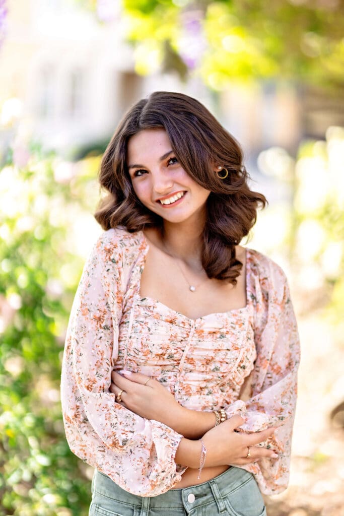 Senior girl with dark hair in a floral top crossing her arms, smiling in front of spring flowers in Downtown Winter Garden.