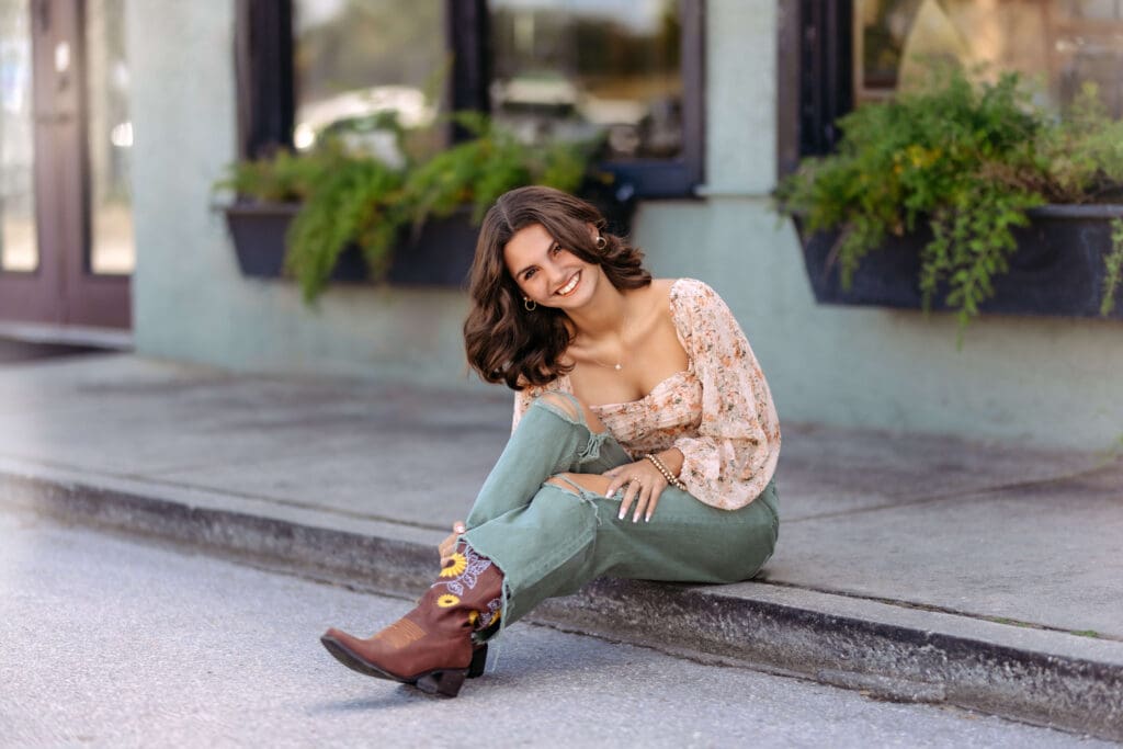 Senior girl in green pants and floral blush top sitting on the curb outside a modern storefront in Downtown Winter Garden. 