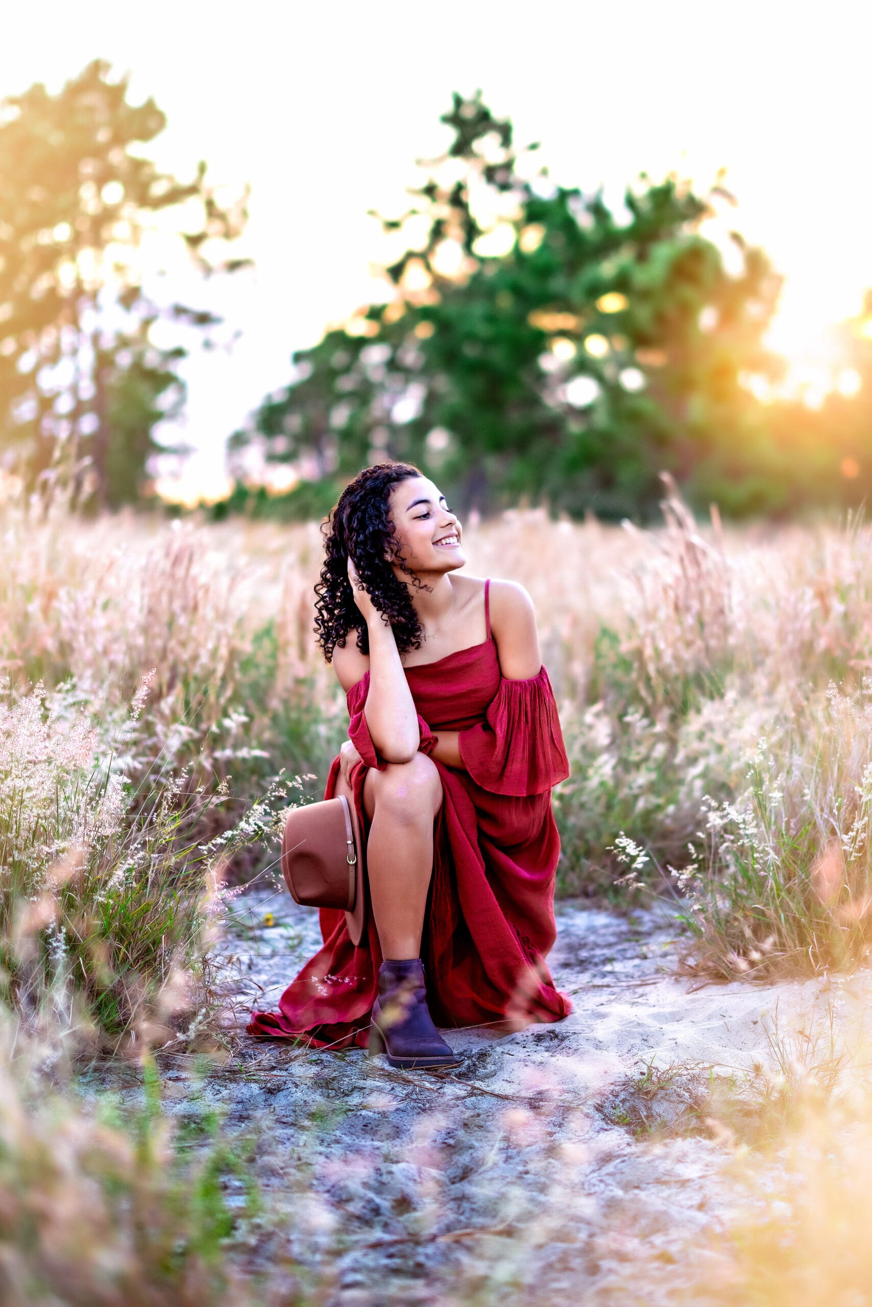 High school senior girl in a deep red dress sitting in a golden field at Lake Louisa State Park near Orlando, Florida during sunset, photographed by Winter Garden senior photographer Petal & Stone.