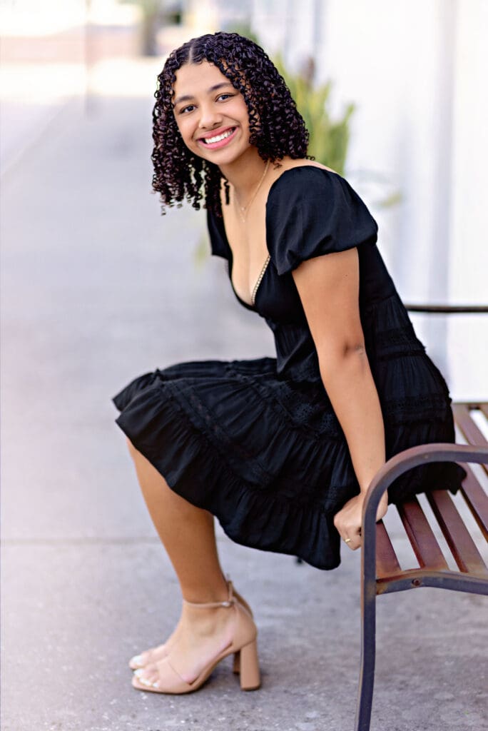 Senior girl in a black dress sitting on a bench with a joyful smile, styled simply and timelessly in Downtown Winter Garden.