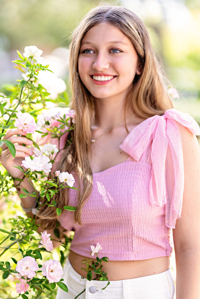 High school girl in a pink shirt with white jeans smiling softly while looking off the the side with gorgeous light pink flower blooms surrounding her in Winter Garden.