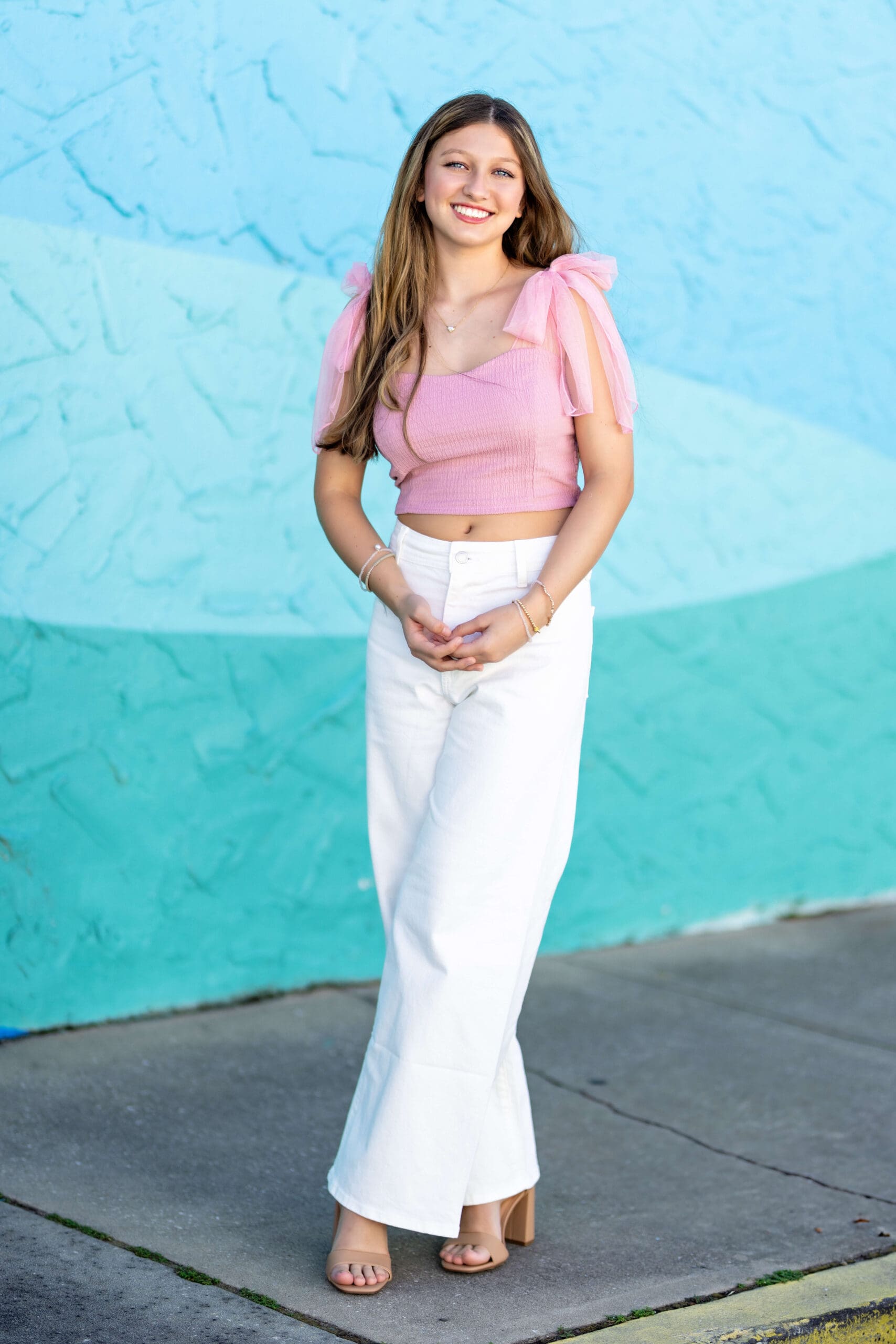 Senior girl in a pink top standing near the sidewalk in front of a teal-blue textured mural wall for senior photos in Downtown Winter Garden.
