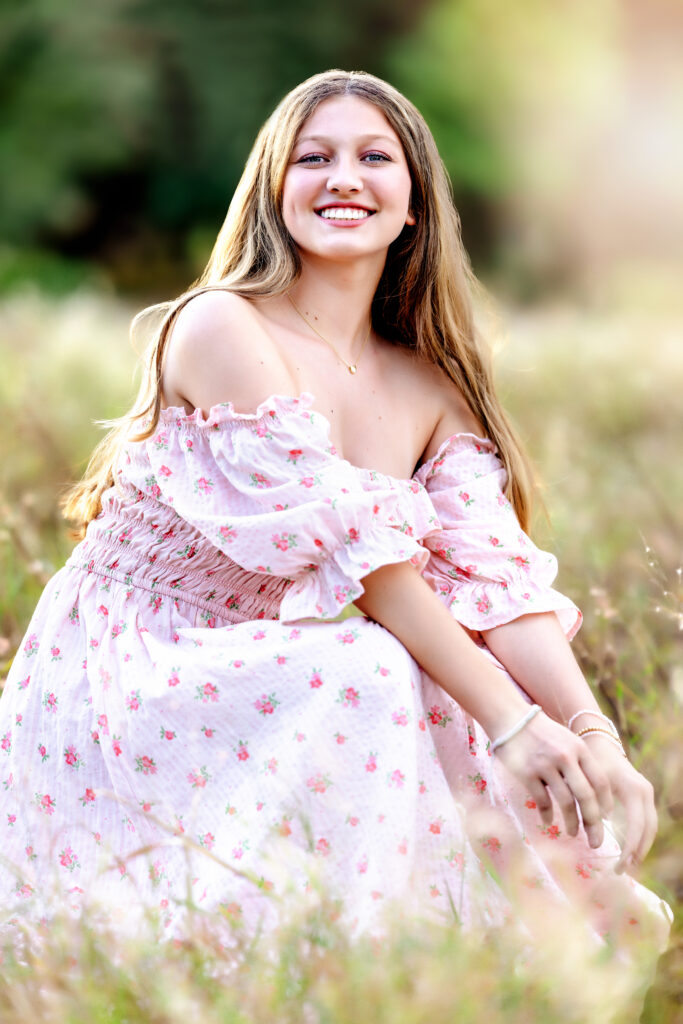 Senior girl laughing while sitting in a golden field, wearing a pink floral dress during a spring photo session in Orlando Florida.