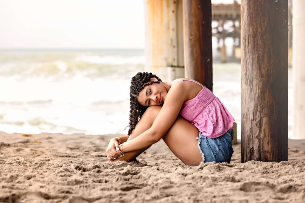 Teen girl curled up under the Cocoa Beach Pier, resting her head on her knees and smiling with beach waves behind her.