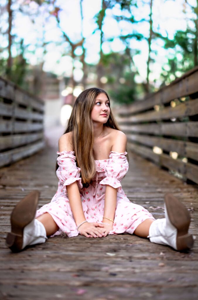 Senior girl in pink floral dress and white boots sitting on a wooden bridge at Lake Louisa State Park, playful and relaxed in a stylish Orlando senior photo session.