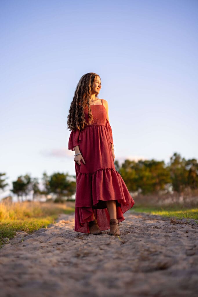 Central Florida senior walking confidently down a sunlit path at Lake Louisa State Park in a flowy red maxi dress, captured in vibrant and bold senior photos.

