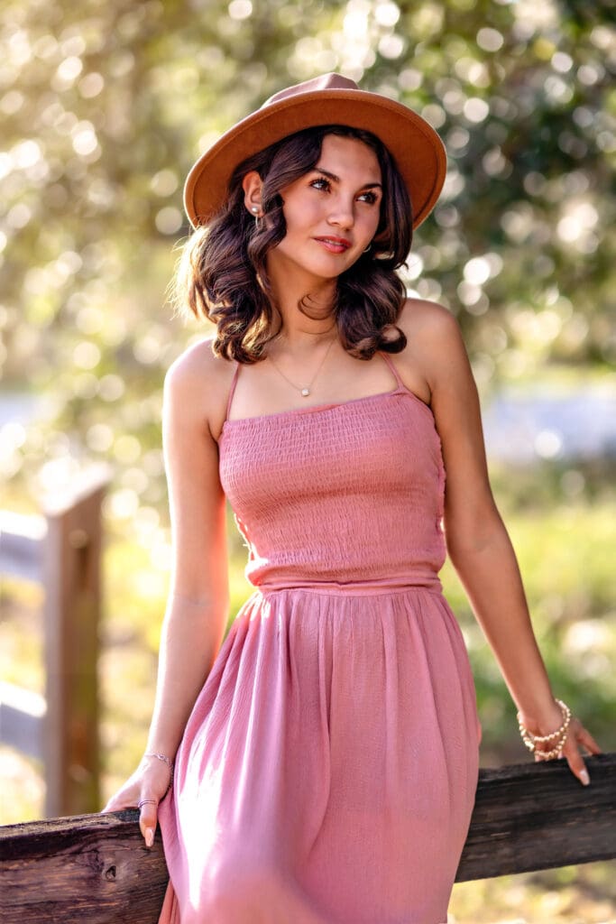 Orlando senior wearing a wide-brimmed hat and rose-colored maxi dress, leaning on a rustic fence at Lake Louisa State Park with golden sunlight creating a dreamy senior photo moment.

