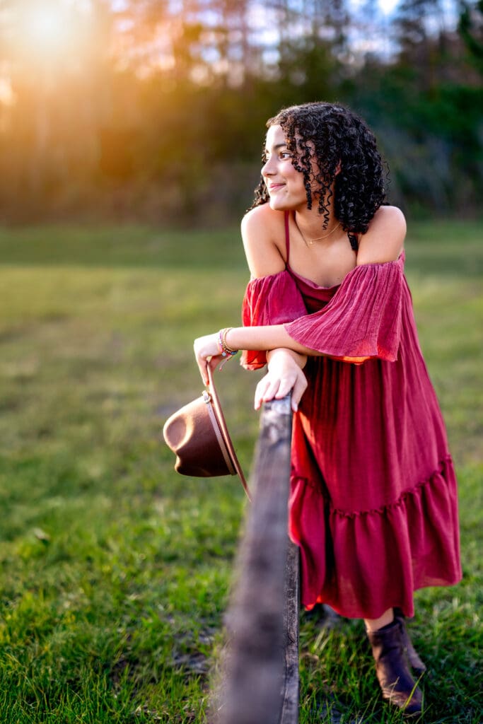 Senior girl in a bold red dress holding a tan hat and leaning on a wooden fence in a grassy field at Lake Louisa State Park, glowing in golden sunset for her Orlando senior photos.

