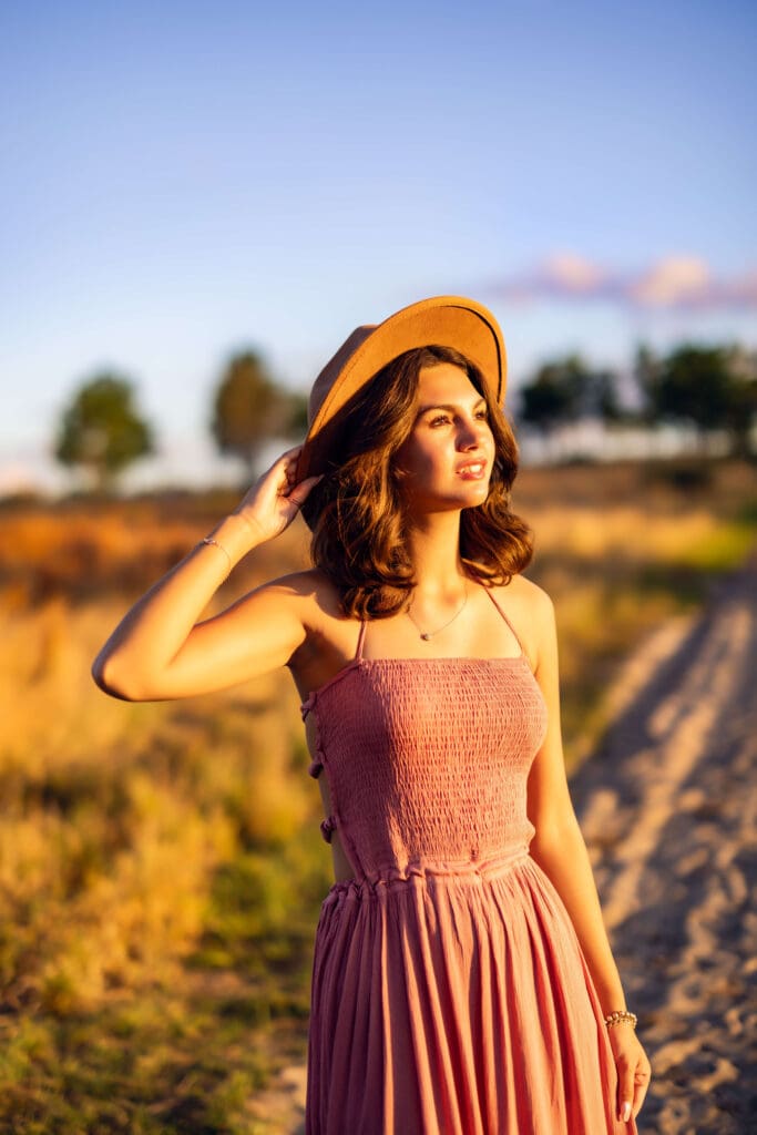 High school senior girl wearing a dusty rose dress and tan hat, bathed in warm sunset light at Lake Louisa State Park, creating a magical golden-hour senior portrait.

