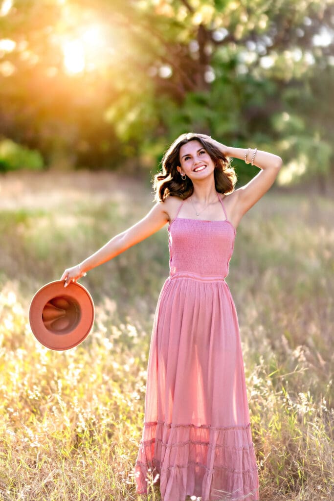 High school senior twirling in a flowy rose-colored maxi dress with a sun hat at Lake Louisa State Park, surrounded by golden hour light and tall grass in a dreamy senior portrait.