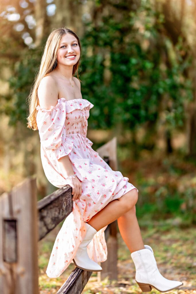 High school senior girl in a pink floral off-shoulder dress sitting on a wooden fence at Lake Louisa State Park, smiling in golden-hour senior portraits near Orlando, Florida.