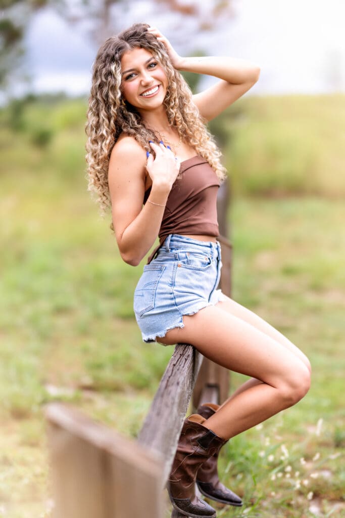 Senior girl sitting on a rustic wooden fence in denim shorts and a brown top during her Orlando, Florida senior photography session.