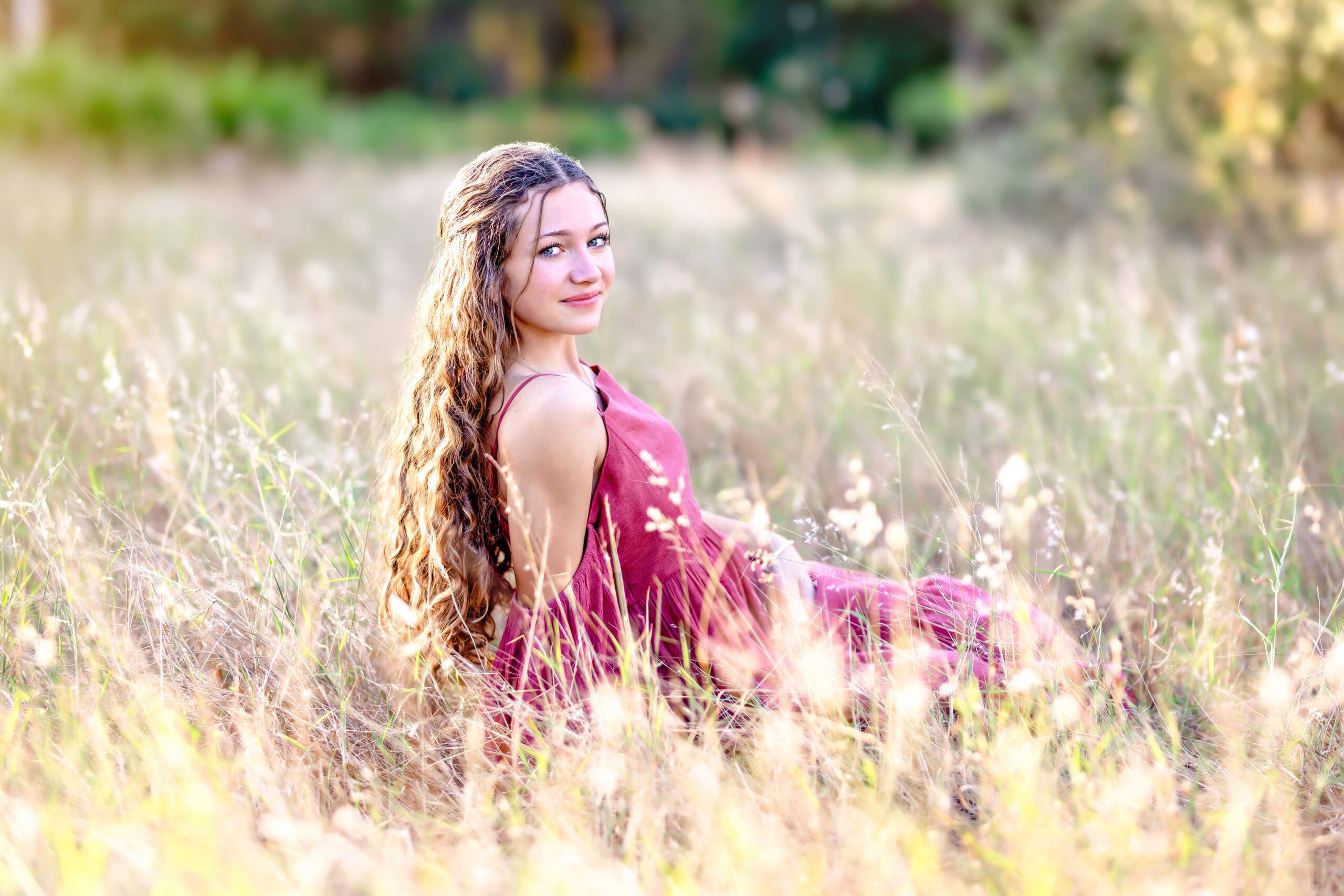 Senior girl in a soft pink dress sitting in tall grass during a golden field senior photo session in Central Florida.