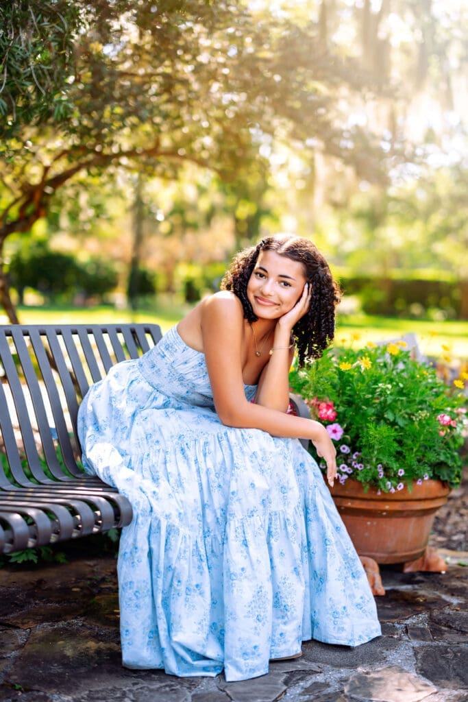 Senior girl in a blue floral dress seated on a garden bench during an outdoor senior portrait session near Orlando Florida